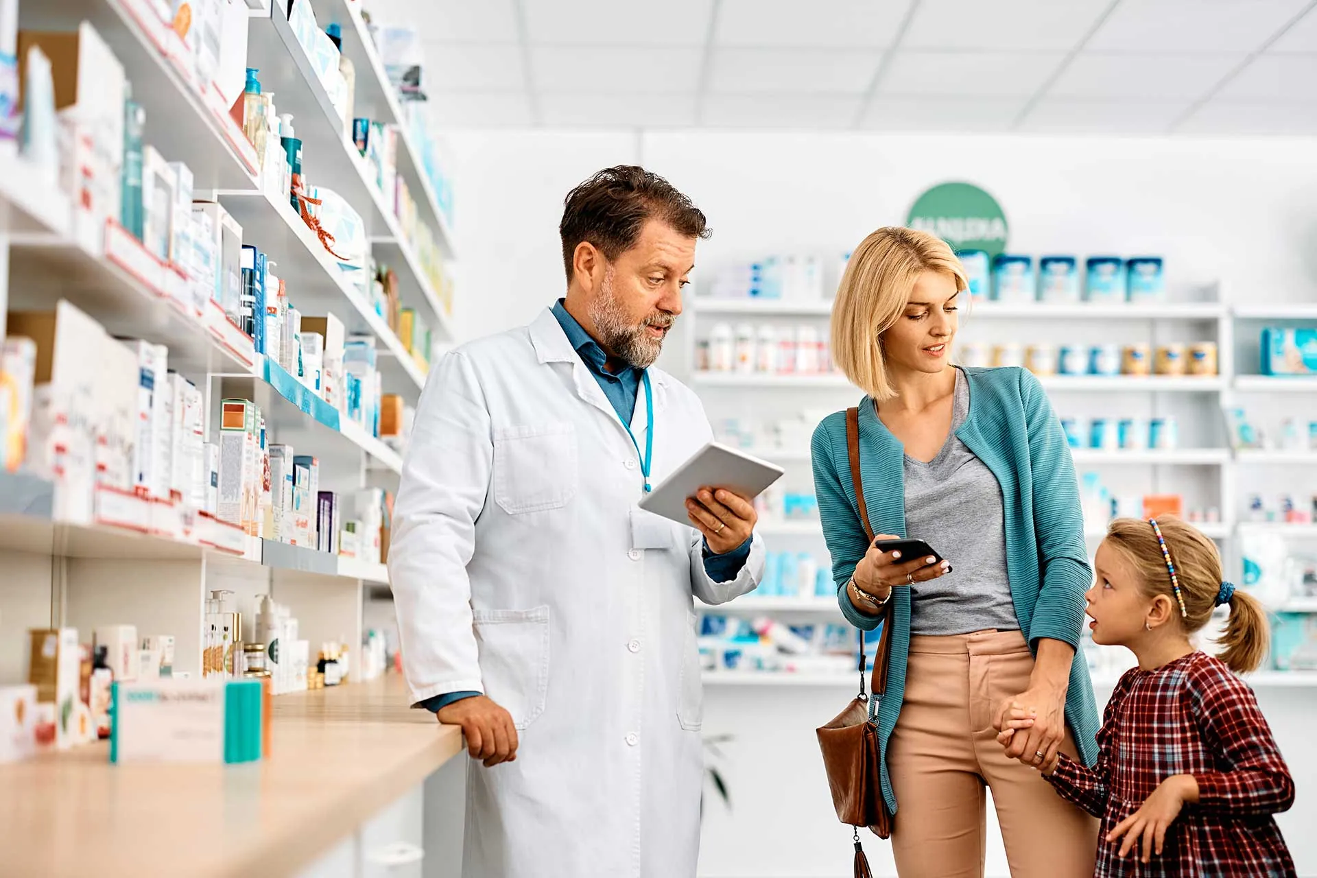 A pharmacist discusses Relief Pharmacy Services with a woman holding a smartphone and a young girl in a pharmacy, surrounded by shelves of medications in the background.