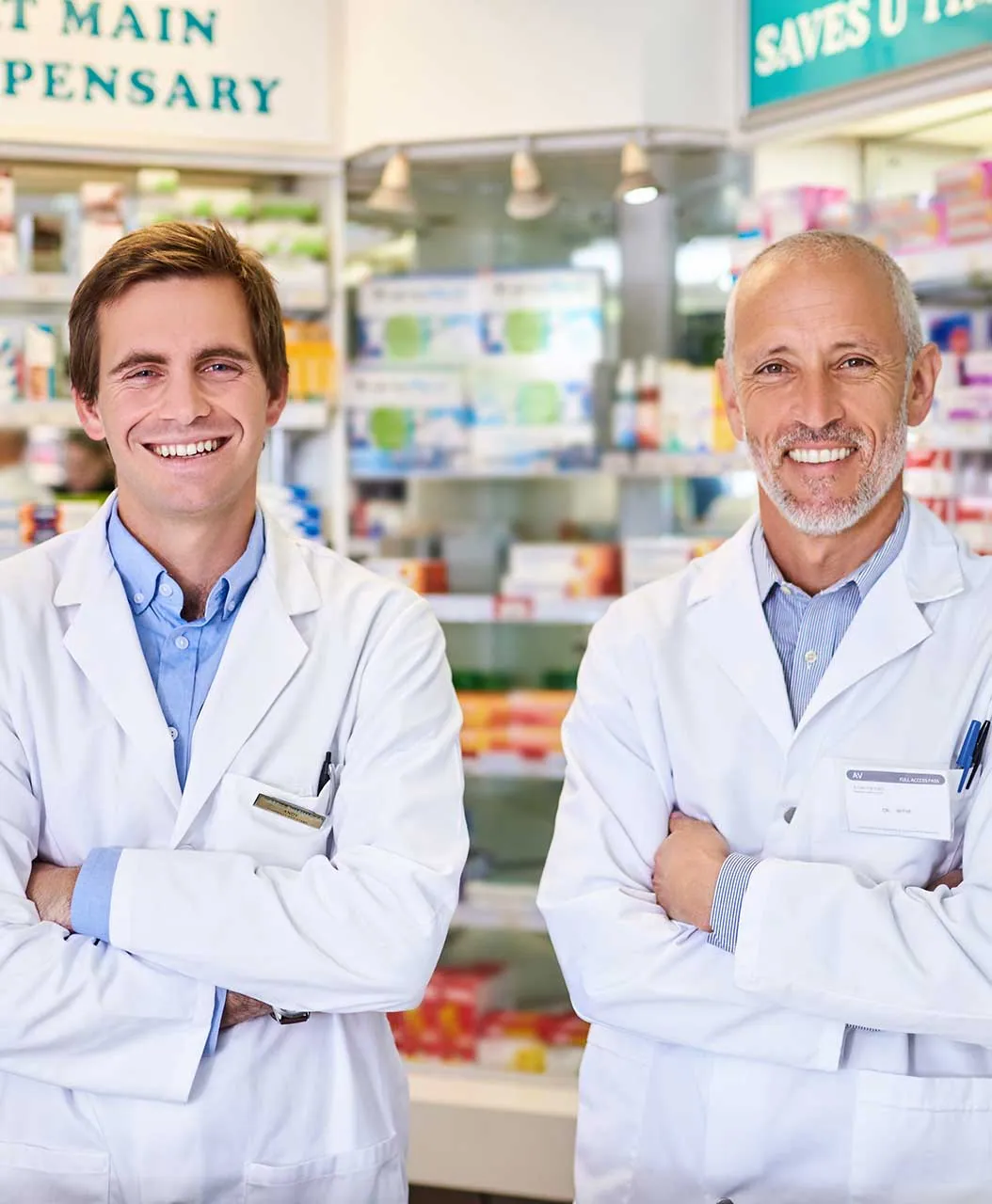 Two male relief pharmacists in white lab coats stand with arms crossed in a pharmacy, shelves of medicine and products visible behind them.