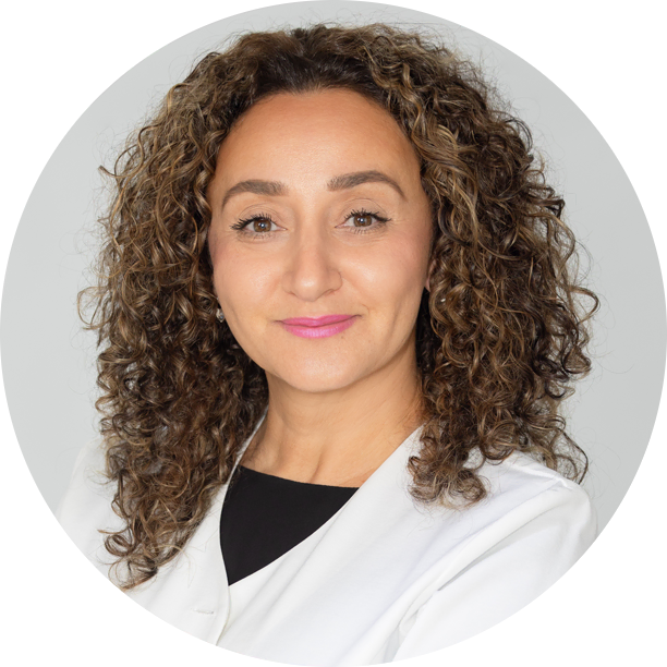 A relief pharmacist with curly brown hair, wearing a white coat and black top, poses for a professional headshot against a light grey background.